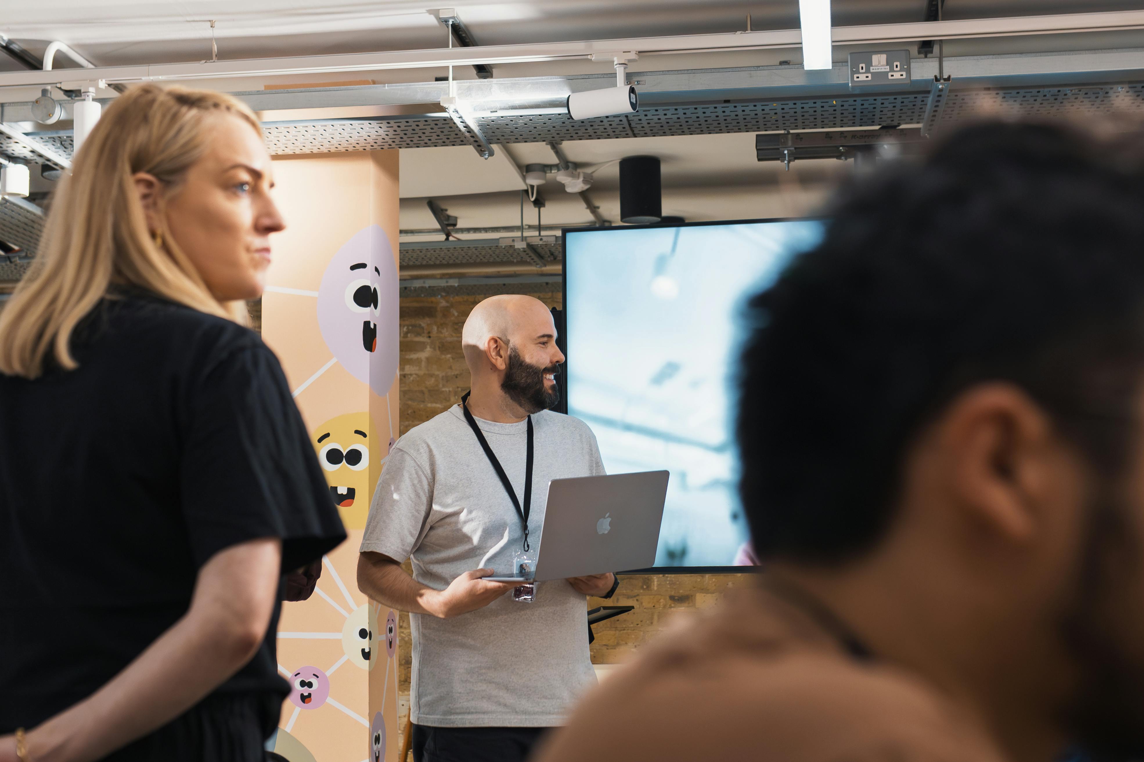 Hombre dando una presentación con una computadora portátil Apple en una sala de capacitación, acompañado de participantes y decoración creativa en el fondo.